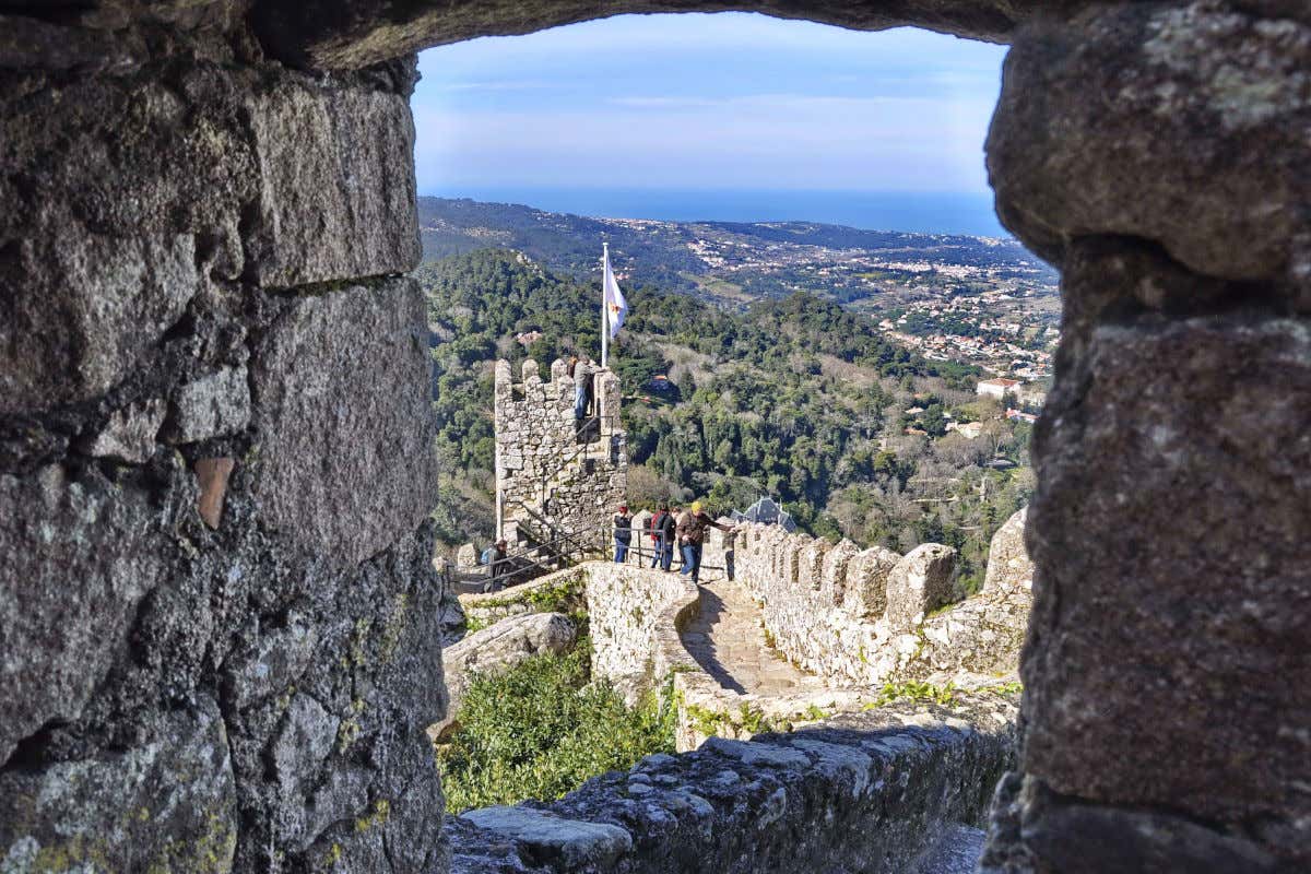 Una ventana de piedra con vistas a los torreones del castelo dos Mouros, un castillo de Sintra rodeado de vegetación y ubicado en lo alto de una colina
