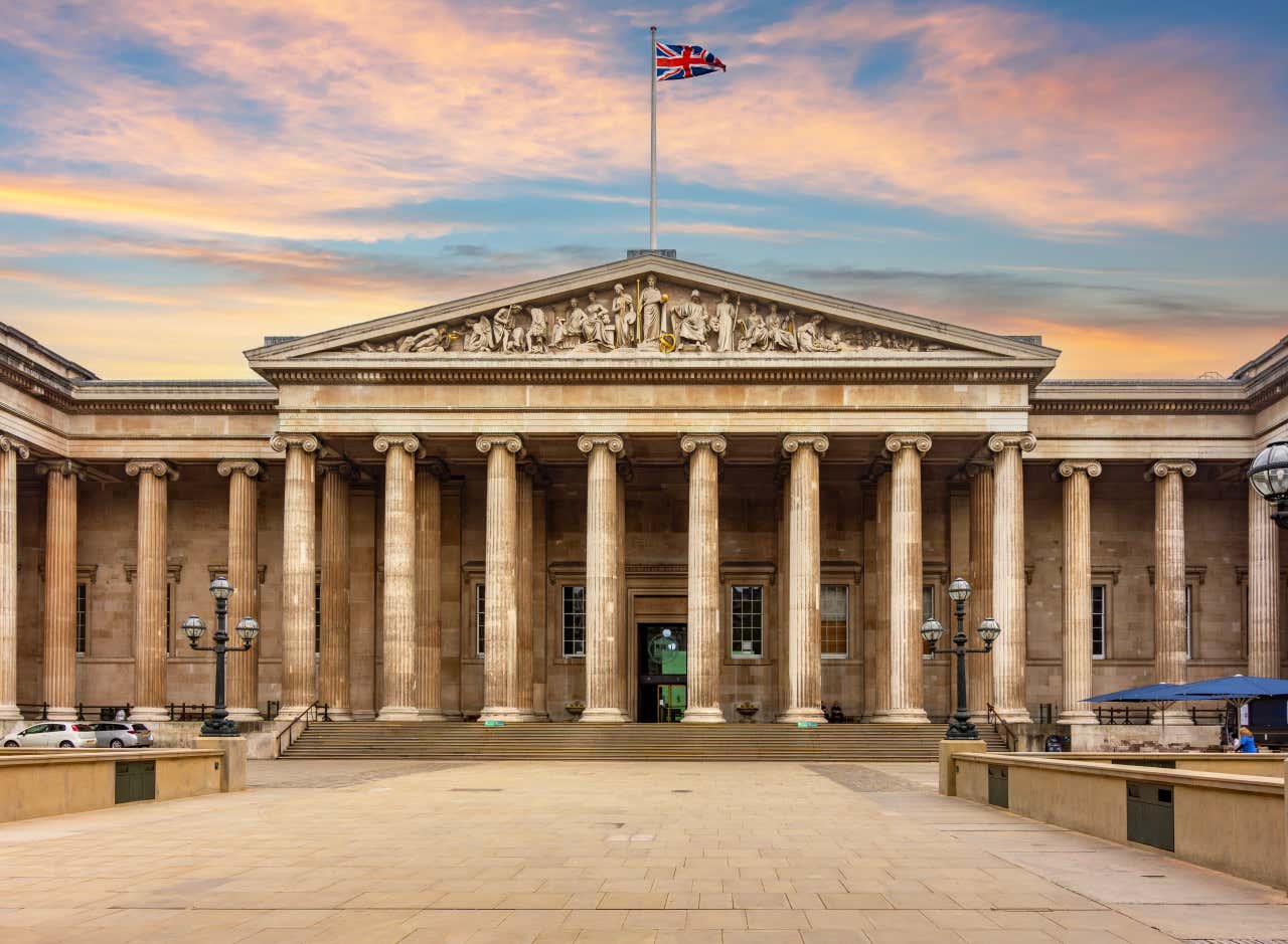 Entrada principal del Museo Británico de Londres, con una bandera de Inglaterra ondeando encima y los colores del atardecer en el cielo