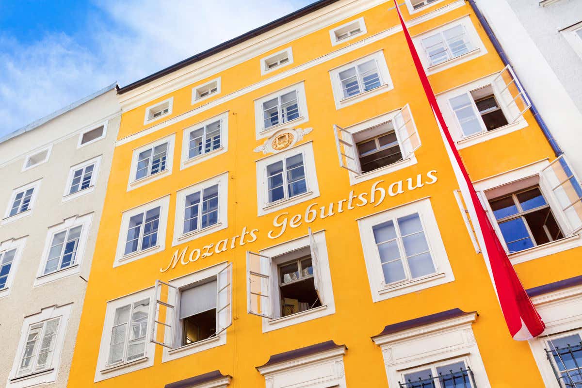 Fachada amarilla de un edificio de unos 5 pisos decorado con una bandera de Austria y unas letras que indican que es la casa natal de Mozart