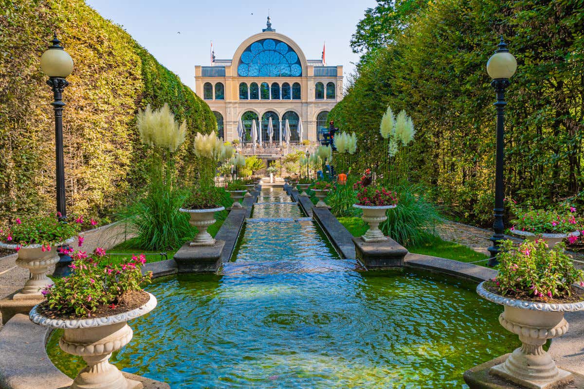 Una fuente rodeada de setos y plantas de camino a un edificio tipo palacete en un jardín botánico