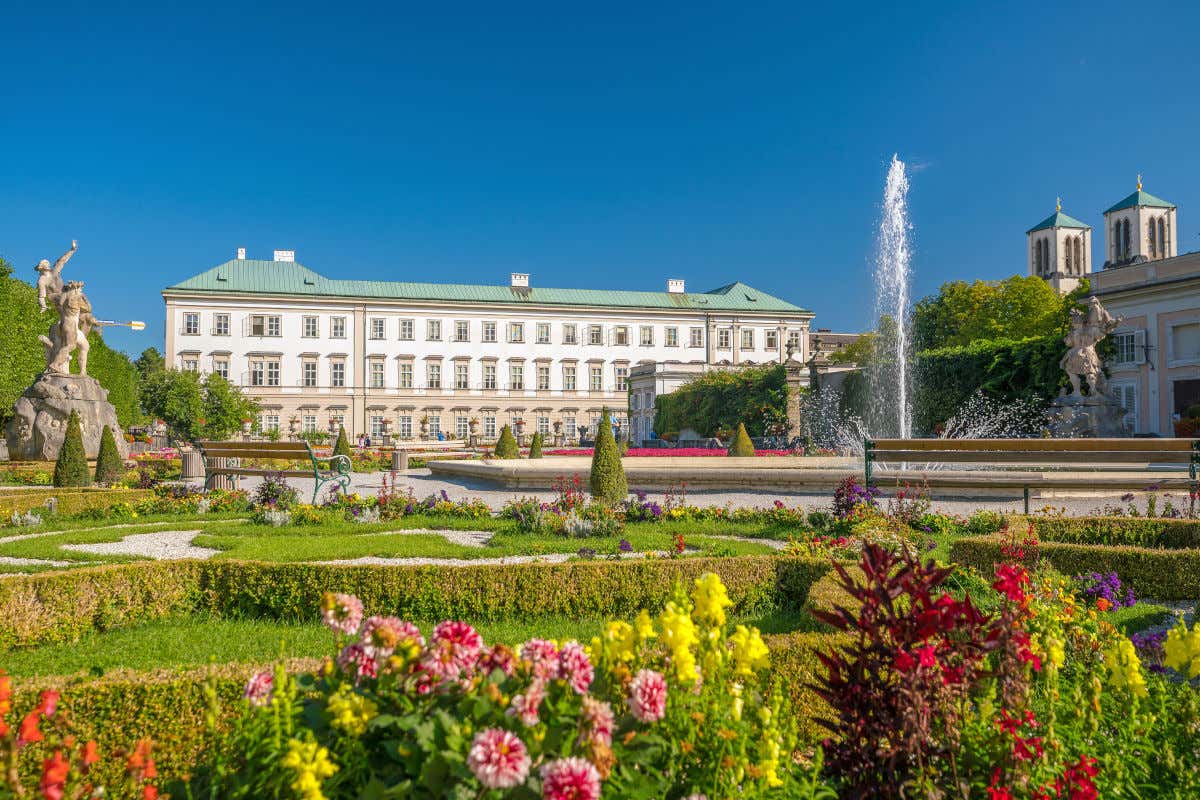 Un palacio de color blanco rodeado de jardines con algunas esculturas y una fuente
