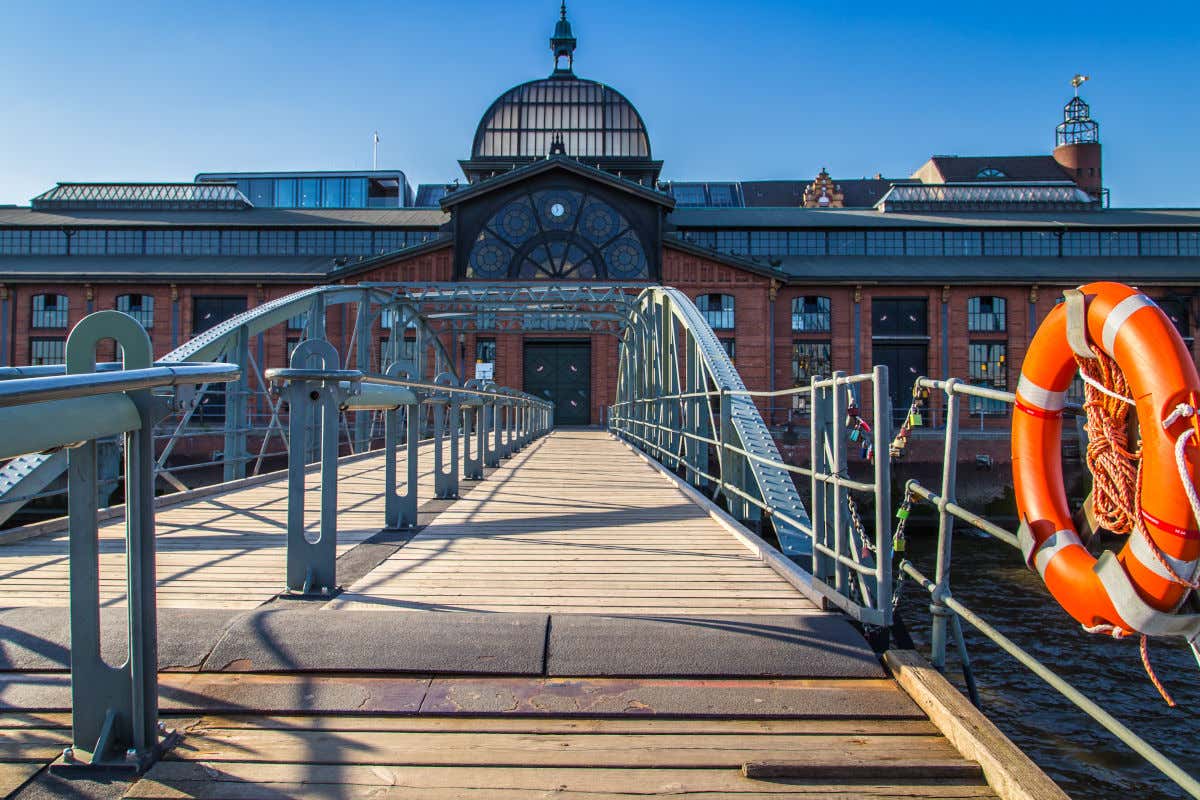 Fachada de un edificio de ladrillo decorado con varias vidrieras y al que se accede a través de un puente que cruza un lago