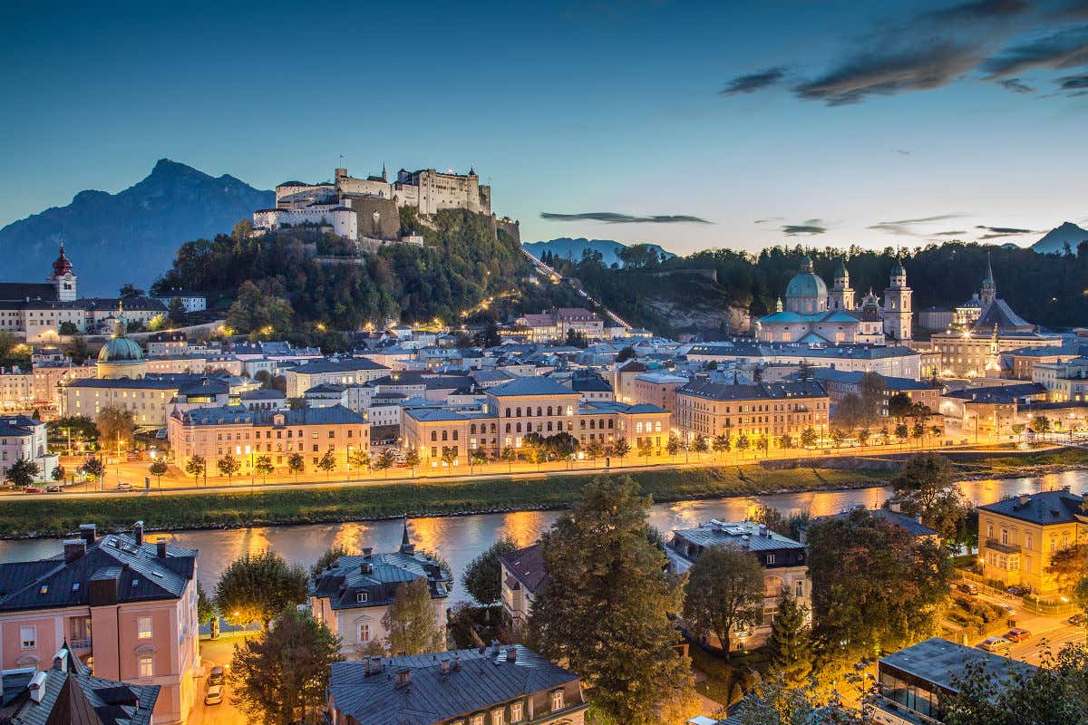 Panorámica de Salzburgo al anochecer con la Catedral, el río y otros edificios iluminados rodeando una colina donde se alza una gran fortaleza