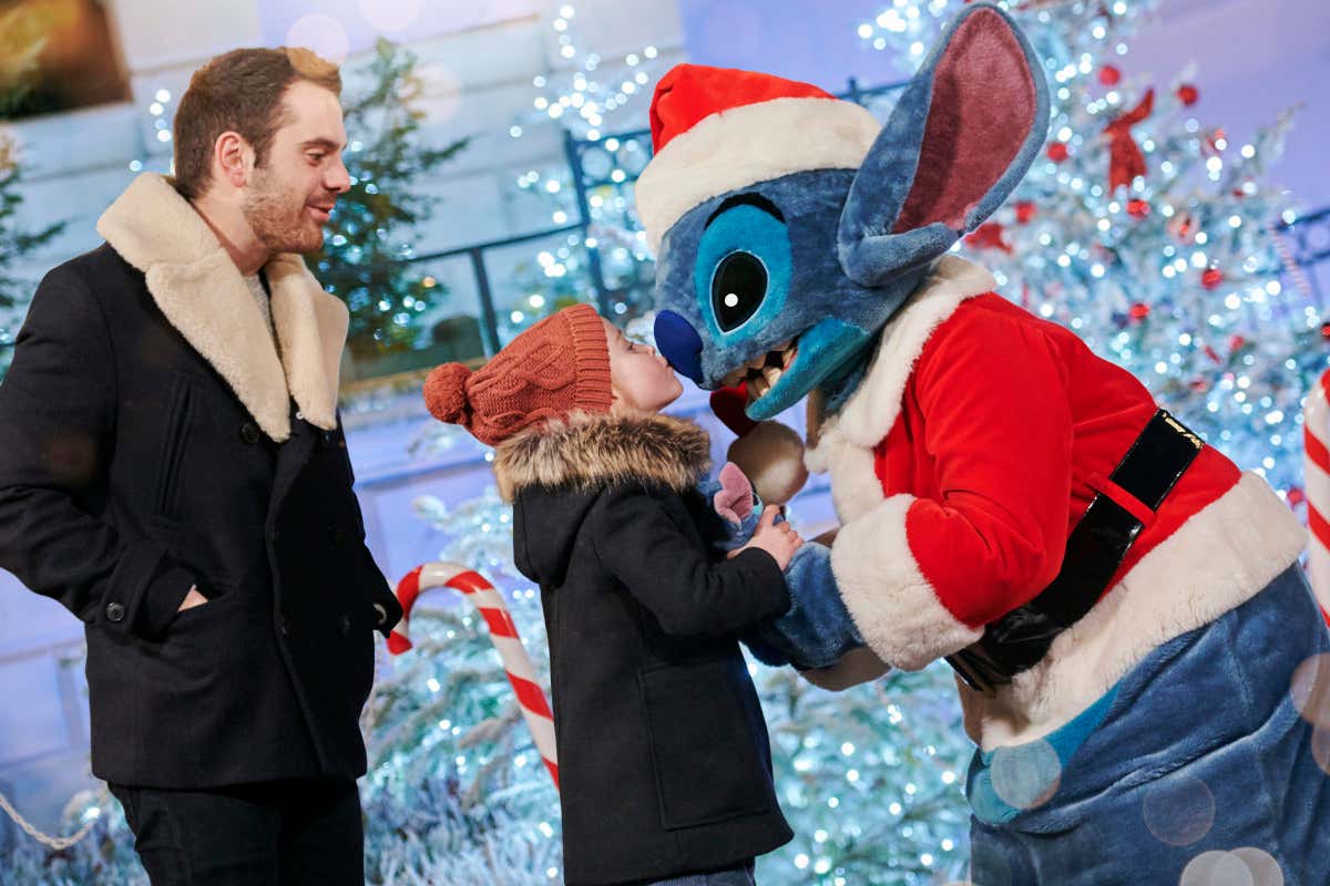 Un niño, bajo la mirada divertida de un hombre, besa y abraza al personaje Stitch de Disney, disfrazado con un traje de Papá Noel
