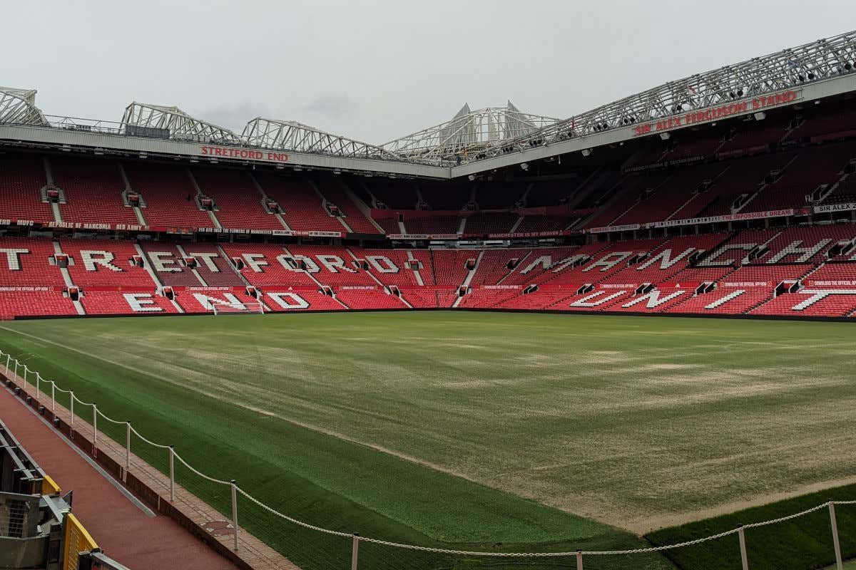 Interior del estadio de fútbol del Manchester United, con el césped sin jugadores y un amplio graderío rojo sin público