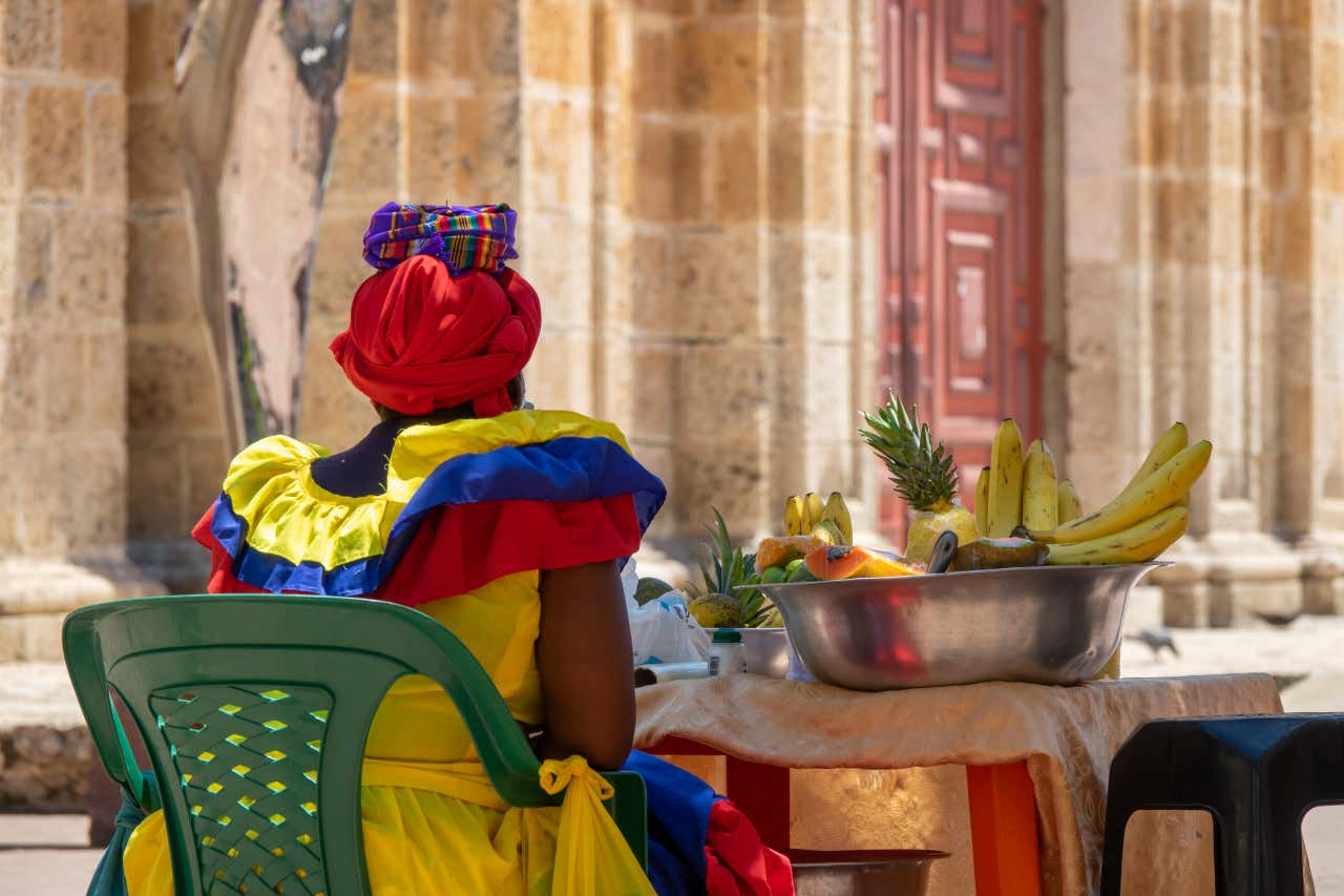 Una mujer vestida con un traje típico colombiano sentada en una plaza con un puesto ambulante de frutas