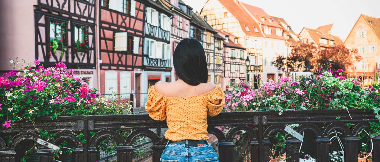 Una mujer de espaldas a la cámara apoyada en un puente con vistas a un canal de Colmar y a varias casas de colores con vigas de madera descubierta