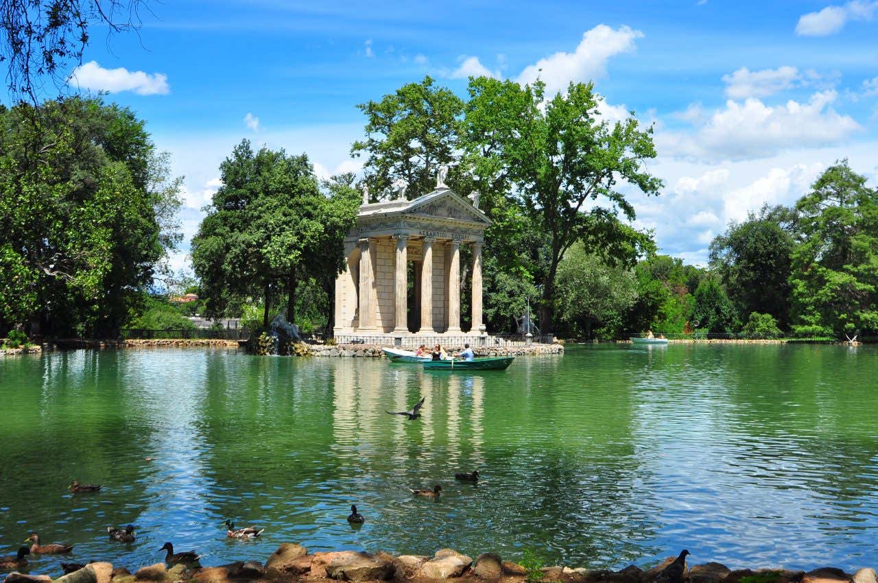 Un tempietto in stile neoclassico in riva a un laghetto immerso nel verde con alcune persone che navigano in barche a remi