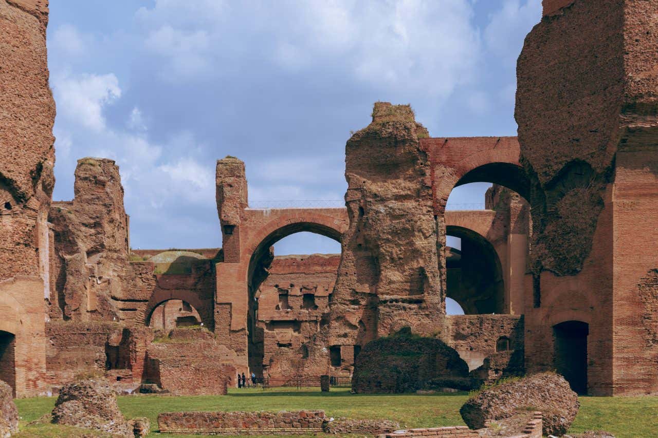 I resti in mattoni rossi delle terme di caracalla, con archi a tutto sesto, mura spoglie e alcuni turisti che passeggiano sull'erba circostante le rovine