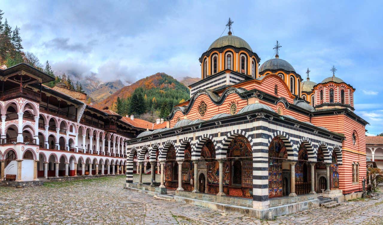 The stunning black and white striped facade of the Rila Monastery in Bulgaria, a complex of ornate Orthodox architecture surrounded by autumn trees.