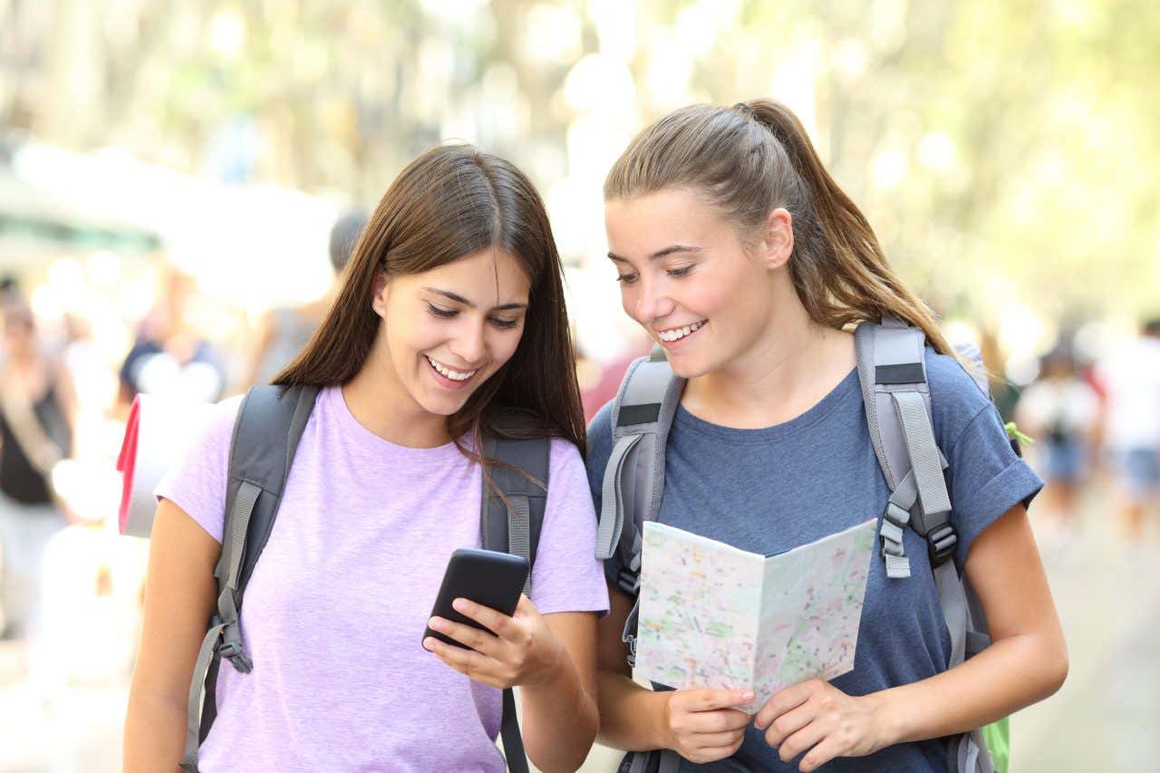 Due giovani donne sorridenti con zaini in spalla osservano una mappa e lo schermo di un cellulare mentre sono in visita in una città europea