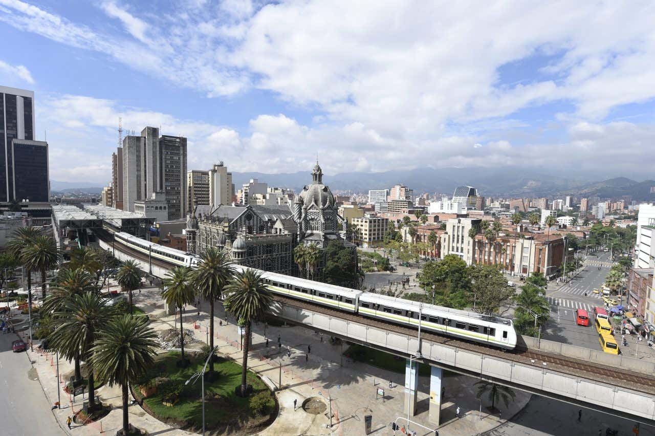 Metro di Medellín su binari sopraelevati che attraversa una piazza urbana con palme, edifici storici e moderni grattacieli visibili sotto un cielo nuvoloso