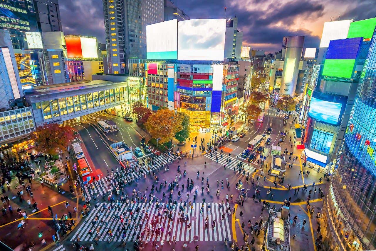 Shibuya Crossing a Tokyo di sera con centinaia di persone che attraversano l'intersezione sotto le luci al neon dei grandi schermi pubblicitari e cartelloni luminosi