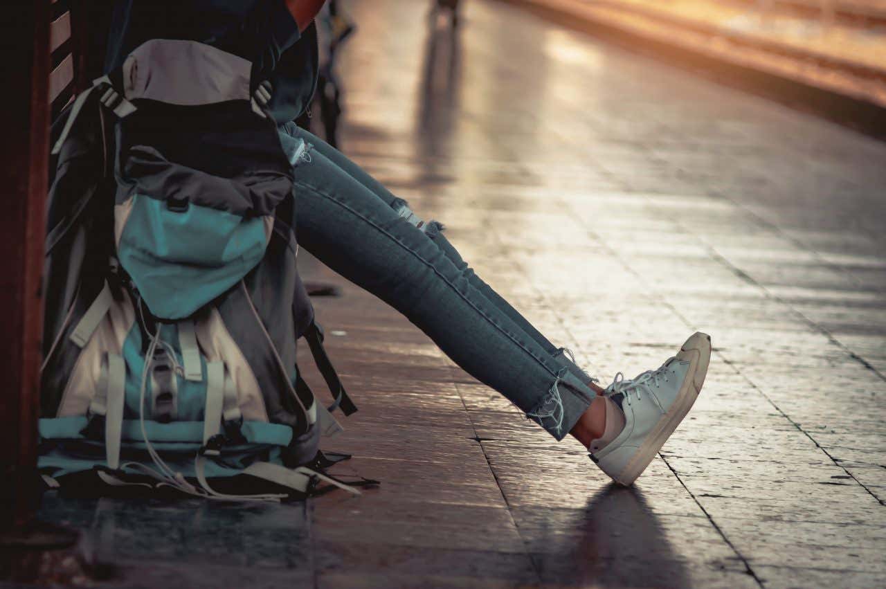 Una persona attende seduta su un grosso zaino da trekking l'arrivo di un treno alla banchina di una stazione ferroviaria