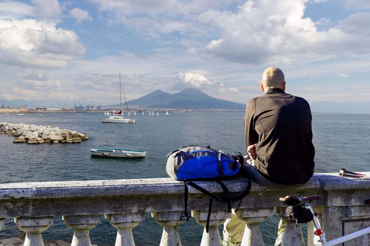 Un uomo seduto su una ringhiera in marmo osserva il Golfo di Napoli con il Vesuvio sullo sfondo