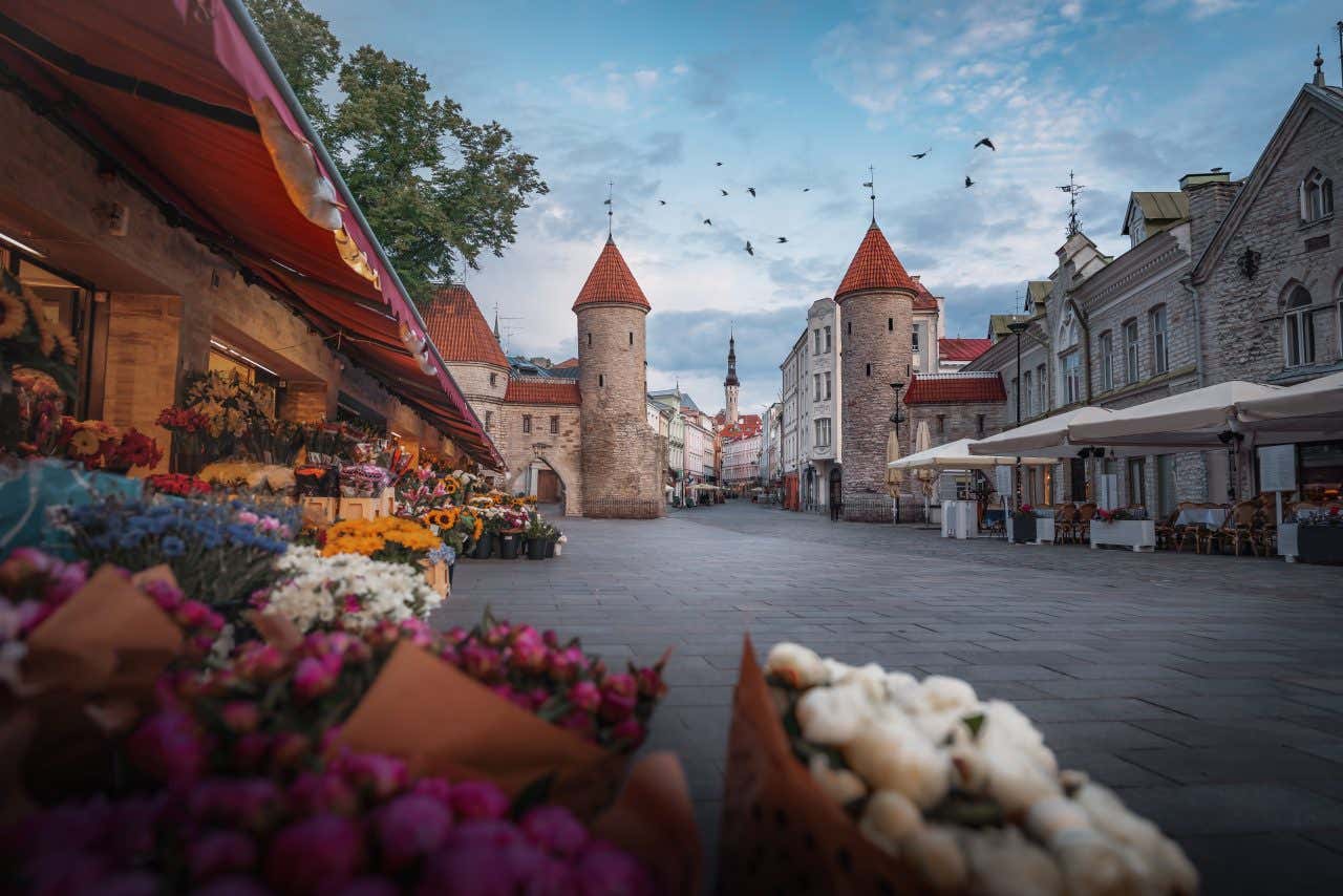 A cobblestone street in the Old Town of Tallinn, Estonia. The foreground features a vibrant flower market with bouquets of various colors. In the background, two medieval stone towers with reddish conical roofs frame the street, with historic buildings lining both sides. Birds are visible flying overhead under a blue and cloudy sky.