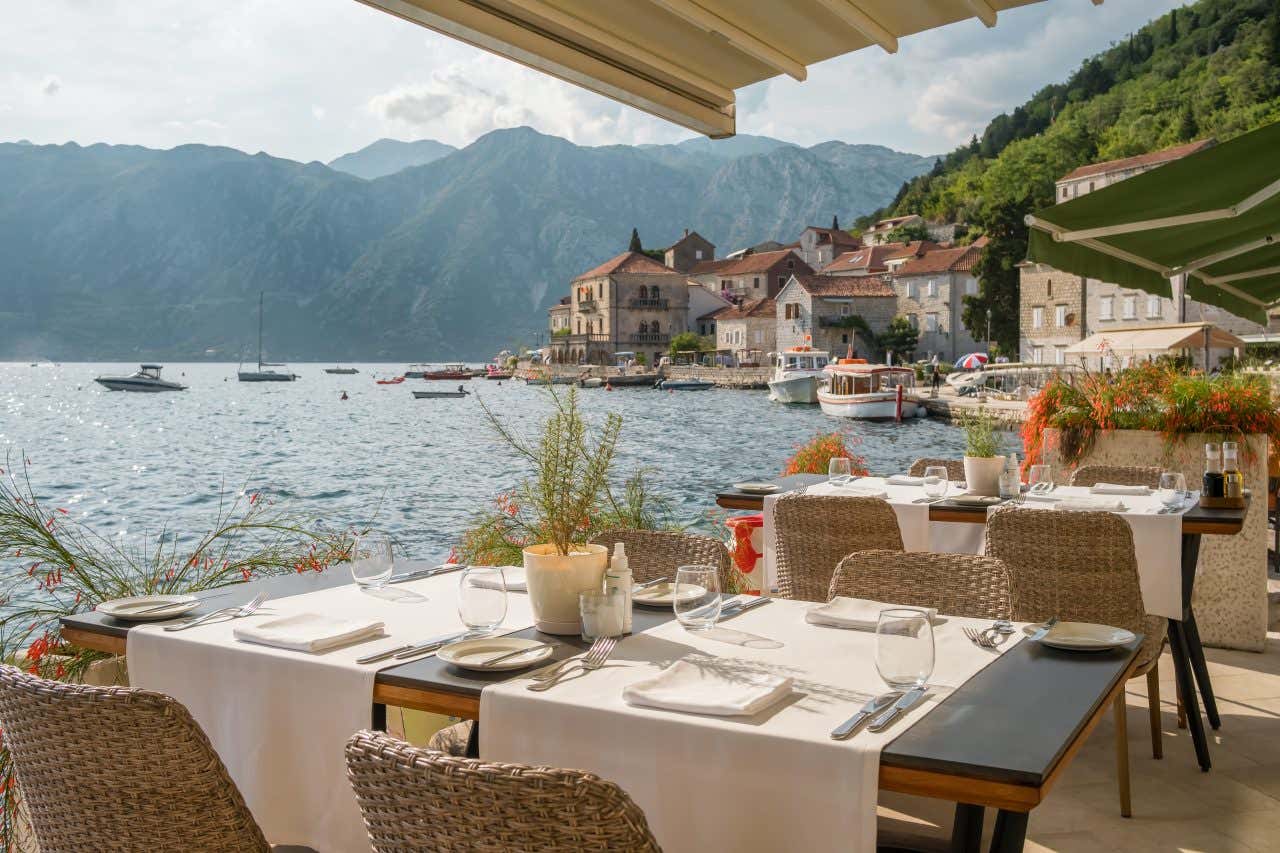 An outdoor restaurant patio overlooking the Bay of Kotor, Montenegro, with set tables and a backdrop of traditional stone houses and green mountains.