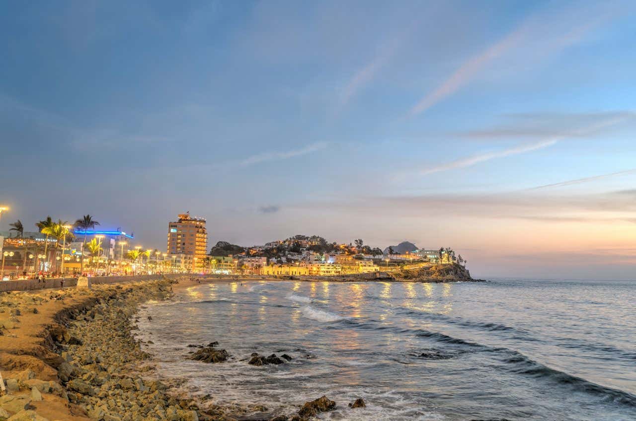 A vibrant seaside promenade in Mazatlán, Mexico, at sunset, with coastal buildings lit up and waves gently crashing on the beach.