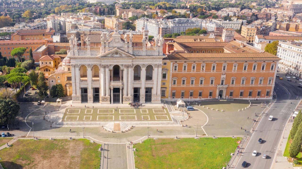Veduta aerea della Basilica di San Giovanni in Laterano a Roma, un’imponente chiesa con facciata bianca in stile barocco, caratterizzata da alte colonne e statue di santi sulla sommità. Davanti si estende una piazza ampia con vialetti geometrici e un prato verde. Intorno si vedono strade, alberi e numerosi edifici color ocra e arancio, tipici dell’architettura romana, con la città che si estende sullo sfondo