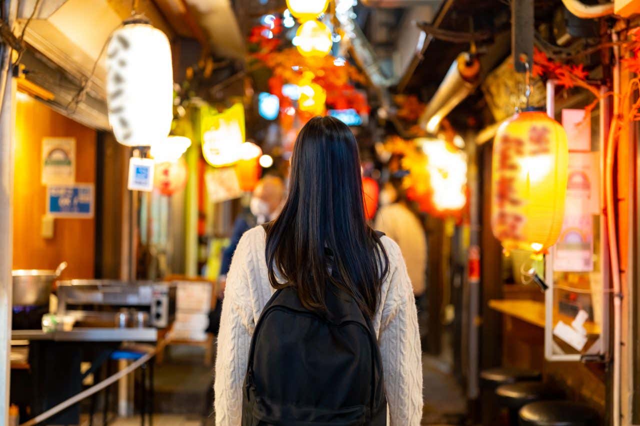 Viaggiatrice vista di spalle esplora un yokocho, vicolo tradizionale giapponese decorato con lanterne rosse, gialle e bianche e insegne luminose di izakaya