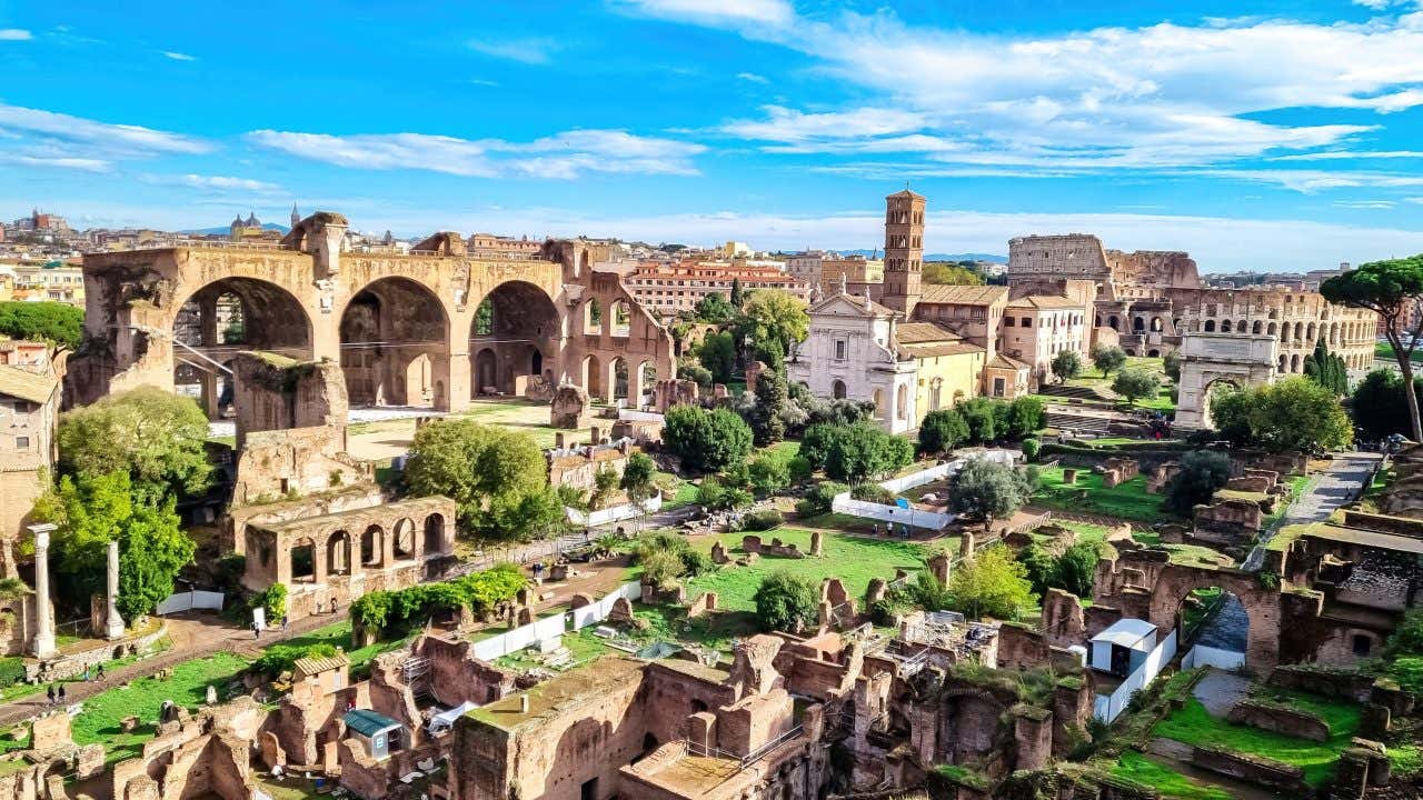 Rovine archeologiche dei Fori Imperiali con colonne antiche e templi romani lungo Via dei Fori Imperiali, Colosseo visibile sullo sfondo sotto cielo azzurro