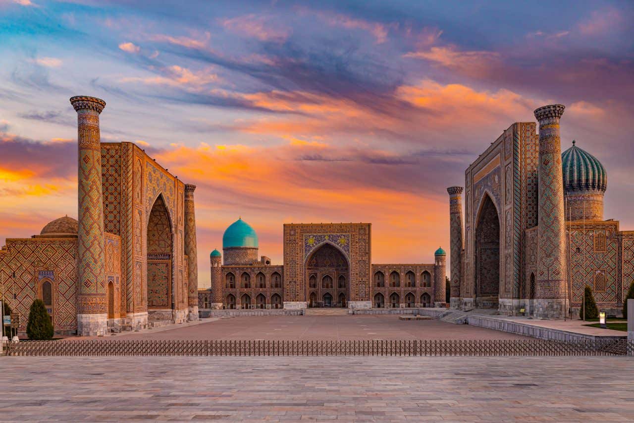 The Registan Square in Samarkand, Uzbekistan, at sunset. The central square is flanked by the Ulugbek Madrasah (left) and the Sher-Dor Madrasah (right), both featuring ornate tilework and tall minarets. The Tilya-Kori Madrasah, with its prominent turquoise dome, is visible in the center background. The sky is filled with vibrant orange and purple clouds.