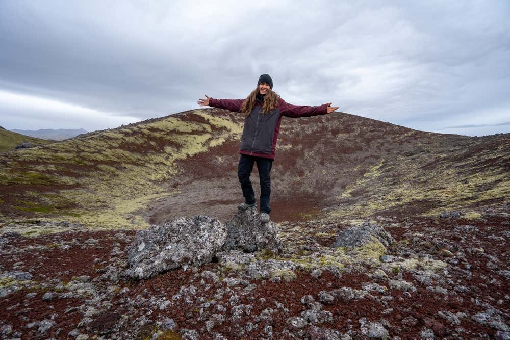Una donna sorridente, con le braccia aperte e in abbigliamento tecnico in posa ai bordi di un cratere di un vulcano islandese