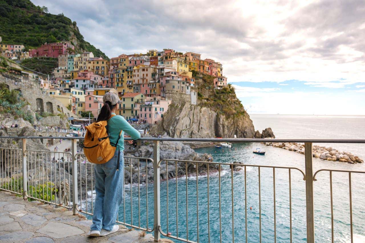 Una ragazza di spalle con una zaino arancione osserva da un punto panoramico un piccolo paese della costa ligure fatto di case colorate arroccate su una scogliera