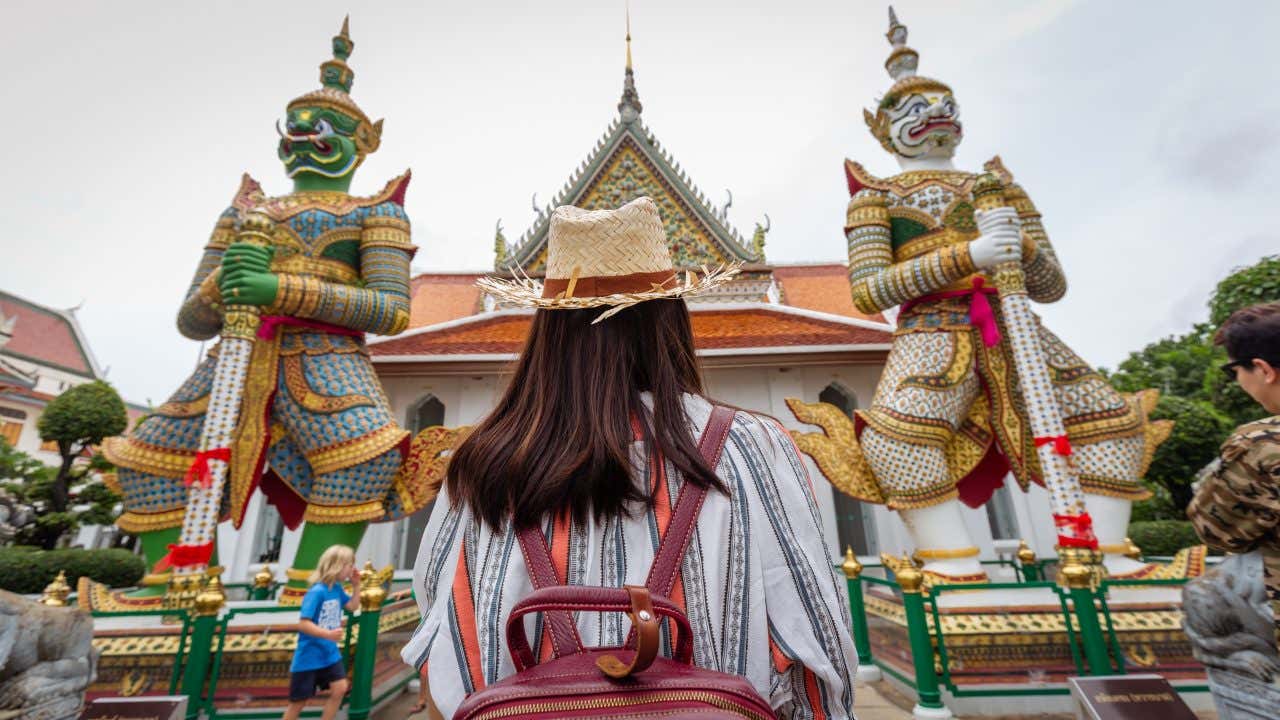 Visitatrice vista di spalle indossa un cappello di paglia e uno zaino bordeaux mentre ammira due monumentali statue di guardiani demoni (Yaksha) riccamente decorati con mosaici colorati, posizionati ai lati dell'ingresso di un tempio thailandese tradizionale con tetto dai tipici ornamenti a punta dorata e tegole arancioni