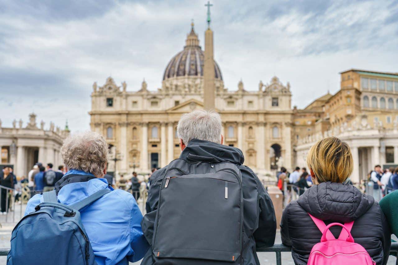 Tre turisti con zaini e giacca a vento di spalle mentre osservano piazza San Pietro in una giornata fredda e nuvolosa