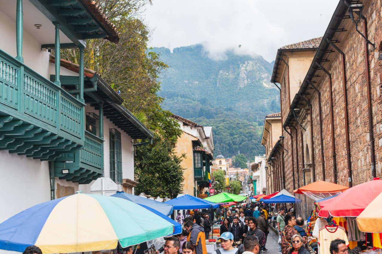 A bustling street market in La Candelaria, Bogotá, Colombia, with colorful umbrellas and vendors beneath a steep, fog-covered mountain.