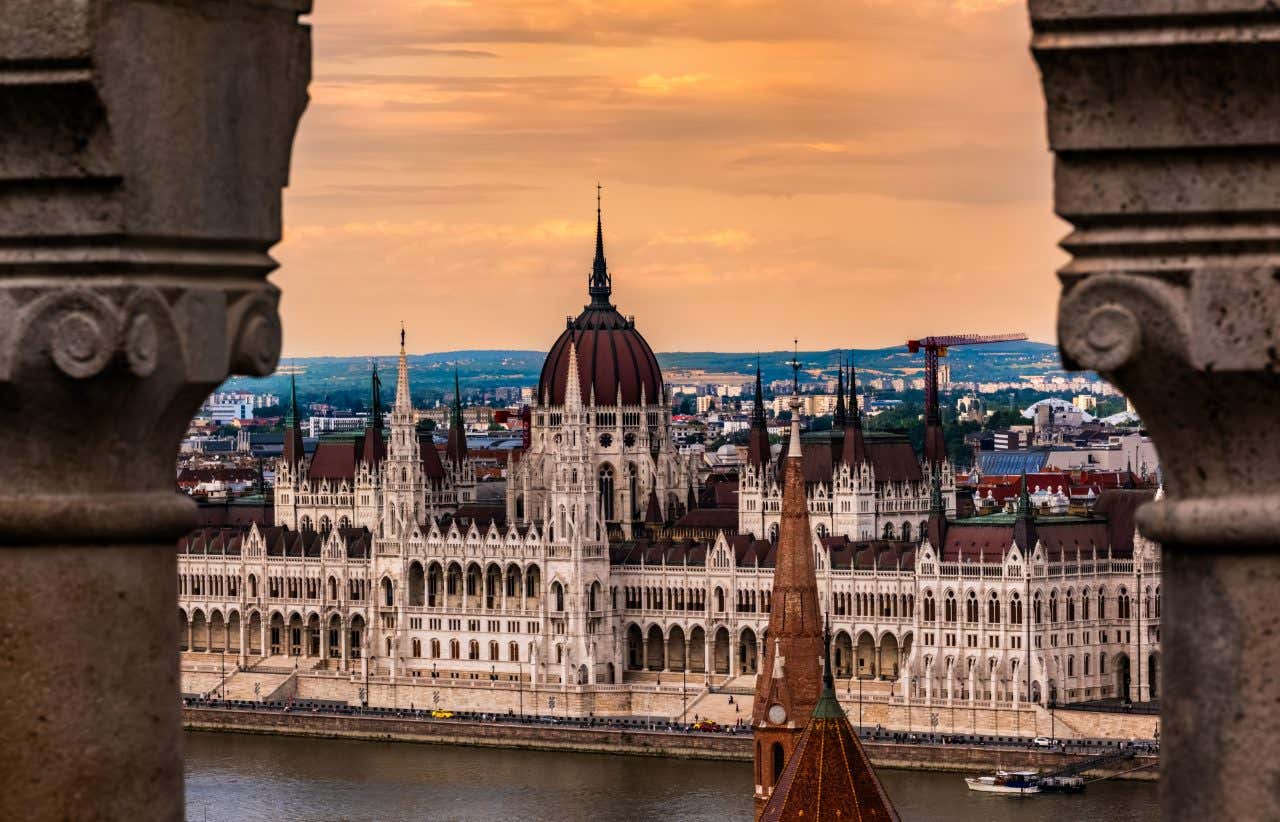 Il maestoso Palazzo del Parlamento Ungherese a Budapest fotografato al tramonto attraverso una cornice architettonica in pietra, con la caratteristica cupola centrale e le facciate neogotiche illuminate dalla luce dorata lungo il fiume Danubio
