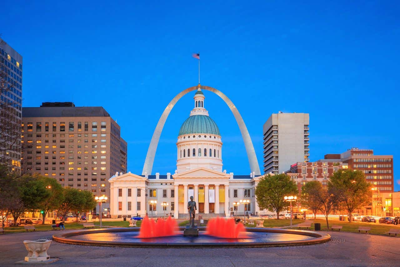 The Old Courthouse and the Gateway Arch in St. Louis, Missouri, at twilight, framed by city buildings and a fountain with red uplighting.