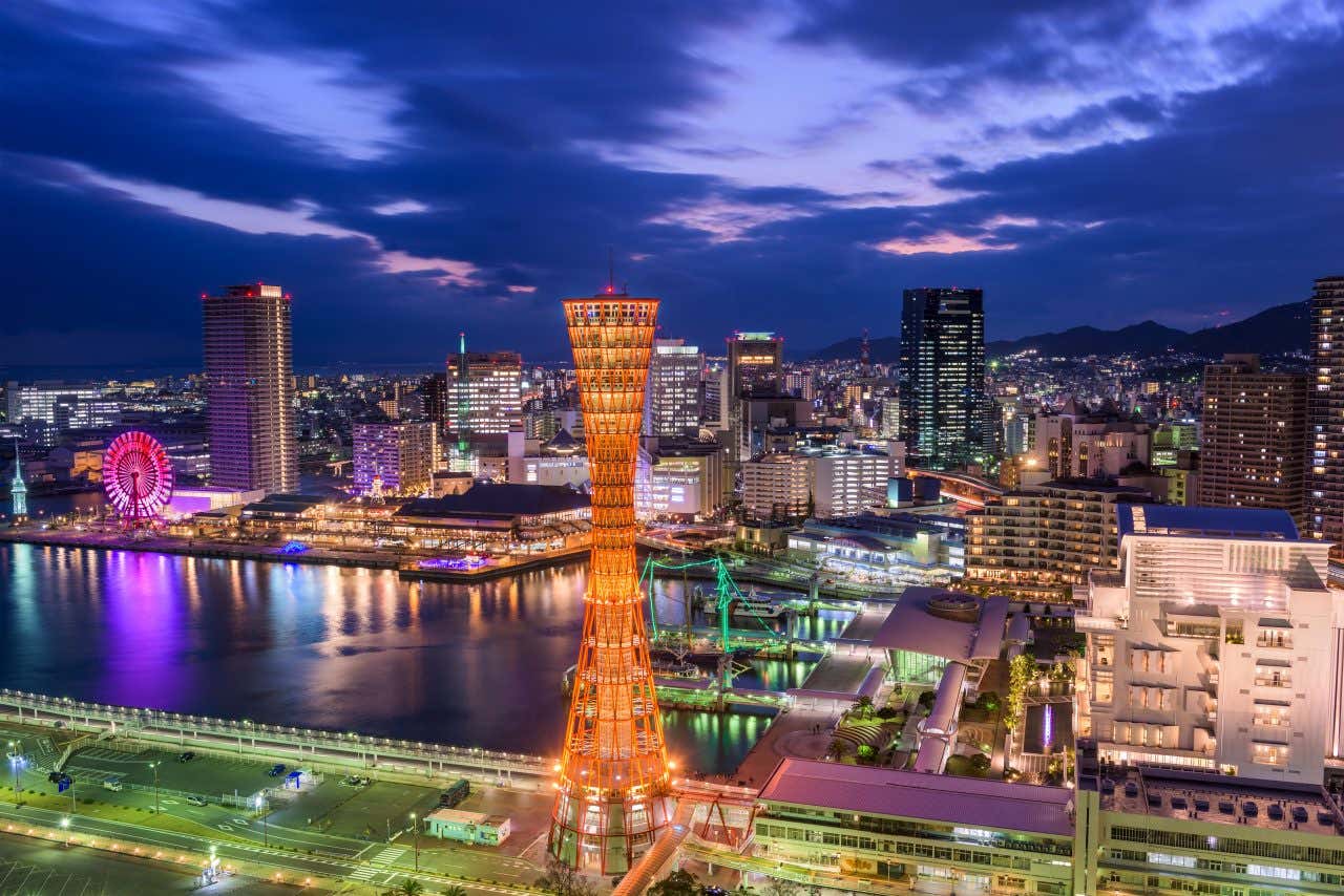 A nighttime skyline view of Kobe, Japan, focusing on the harbor area. The iconic red Kobe Port Tower is illuminated and stands prominently in the center. Modern skyscrapers line the waterfront, and a colorful Ferris wheel is visible to the left. The city lights reflect brightly on the water of the port under a dark blue evening sky.