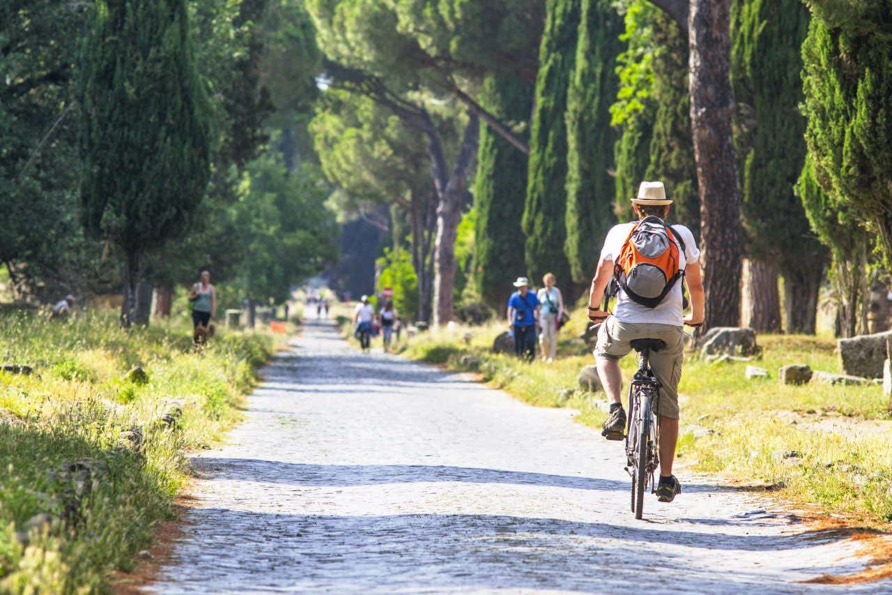 Un uomo in bici pedala lungo una strada immersa nel verde con resti romani a entrambi i lati e altre persone che passeggiano lungo la via Appia