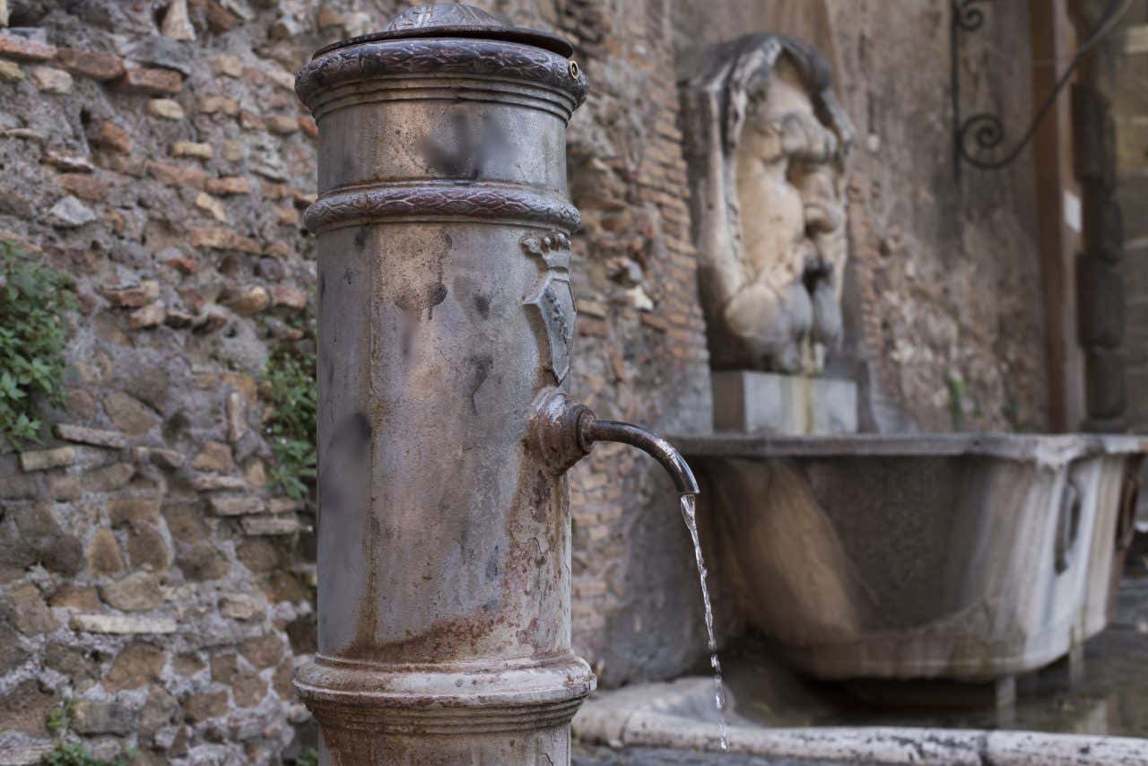 Una fontanella tipica di Roma, di metallo scuro e con il rubinetto curvo e sottile, da cui esce un costante flusso d'acqua