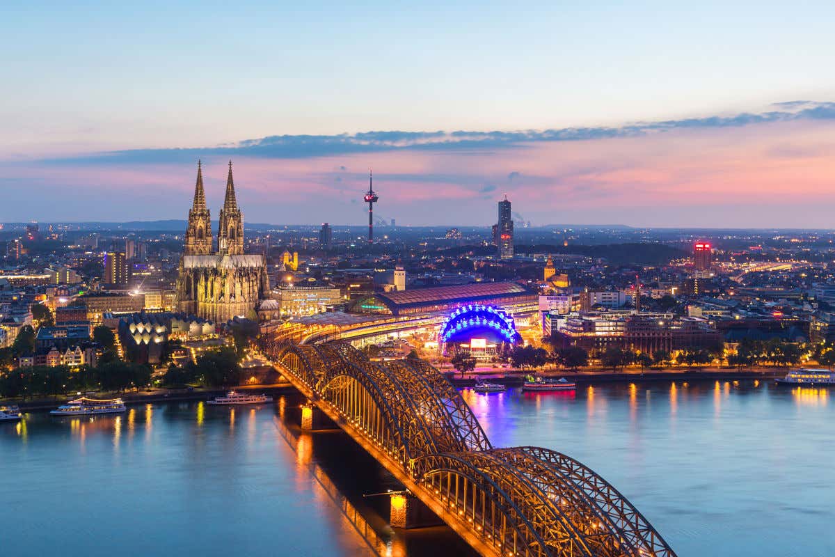 Vista aérea de la ciudad de Colonia al atardecer, con varios barcos navegando por el río Rin y el puente Hohenzollern y la Catedral iluminados
