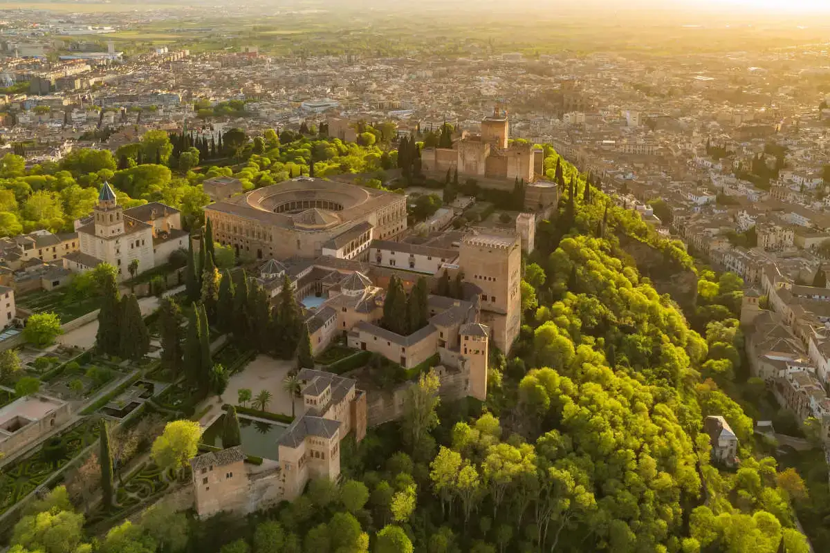 Vista aerea dell'Alhambra di Granada circondata da vegetazione e il resto della città sullo sfondo al tramonto