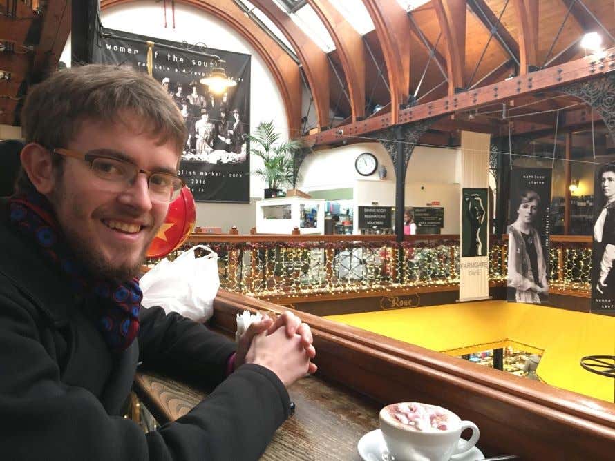 A man sitting at a wooden table drinking a coffee drink in a room with a high ceiling.