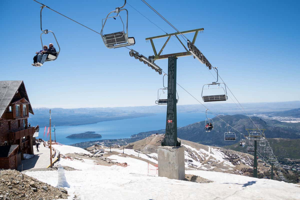Varias personas utilizando un telesilla que sube por una montaña dejando atrás las vistas de un lago y varios cerros en un día despejado