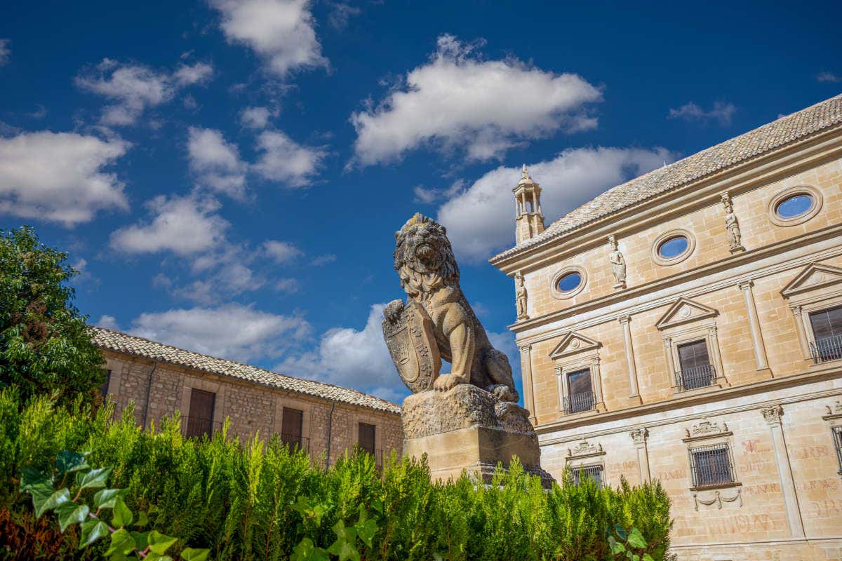 Escultura de piedra de un león sujetando un escudo en una plaza ajardinada rodeada de palacios