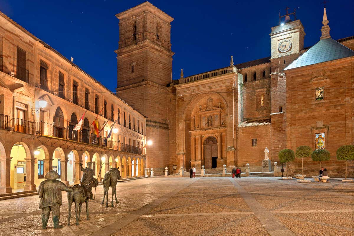 Vistas de una iglesia románica junto a soportales manchegos en una plaza con la escultura de Don Quijote y Sancho Panza