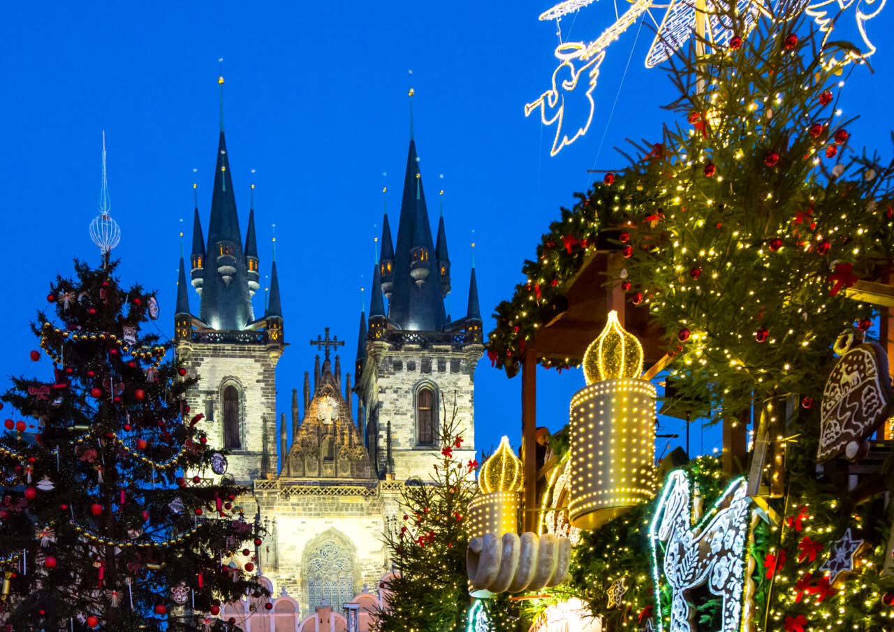 Mercatini di Natale nella piazza della Città Vecchia di Praga con la Chiesa di Tyn illuminata sullo sfondo, alberi di Natale decorati e luminarie dorate al crepuscolo
