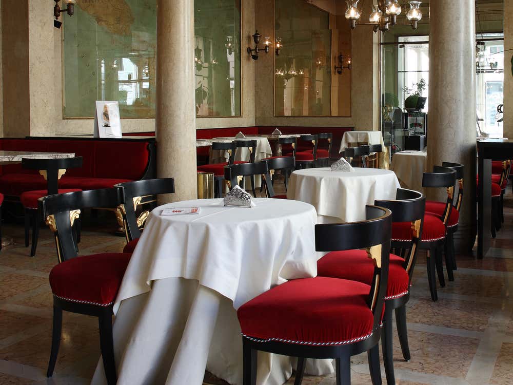 Interno elegante della Sala Verde del Caffè Pedrocchi: sedute imbottite di velluto rosso scuro, tavolini bassi in marmo, pareti chiare con decorazioni classiche, grandi finestre che lasciano entrare luce naturale, lampadari ornamentali appesi al soffitto e specchio sopra un camino che conferisce un’atmosfera raffinata e accogliente