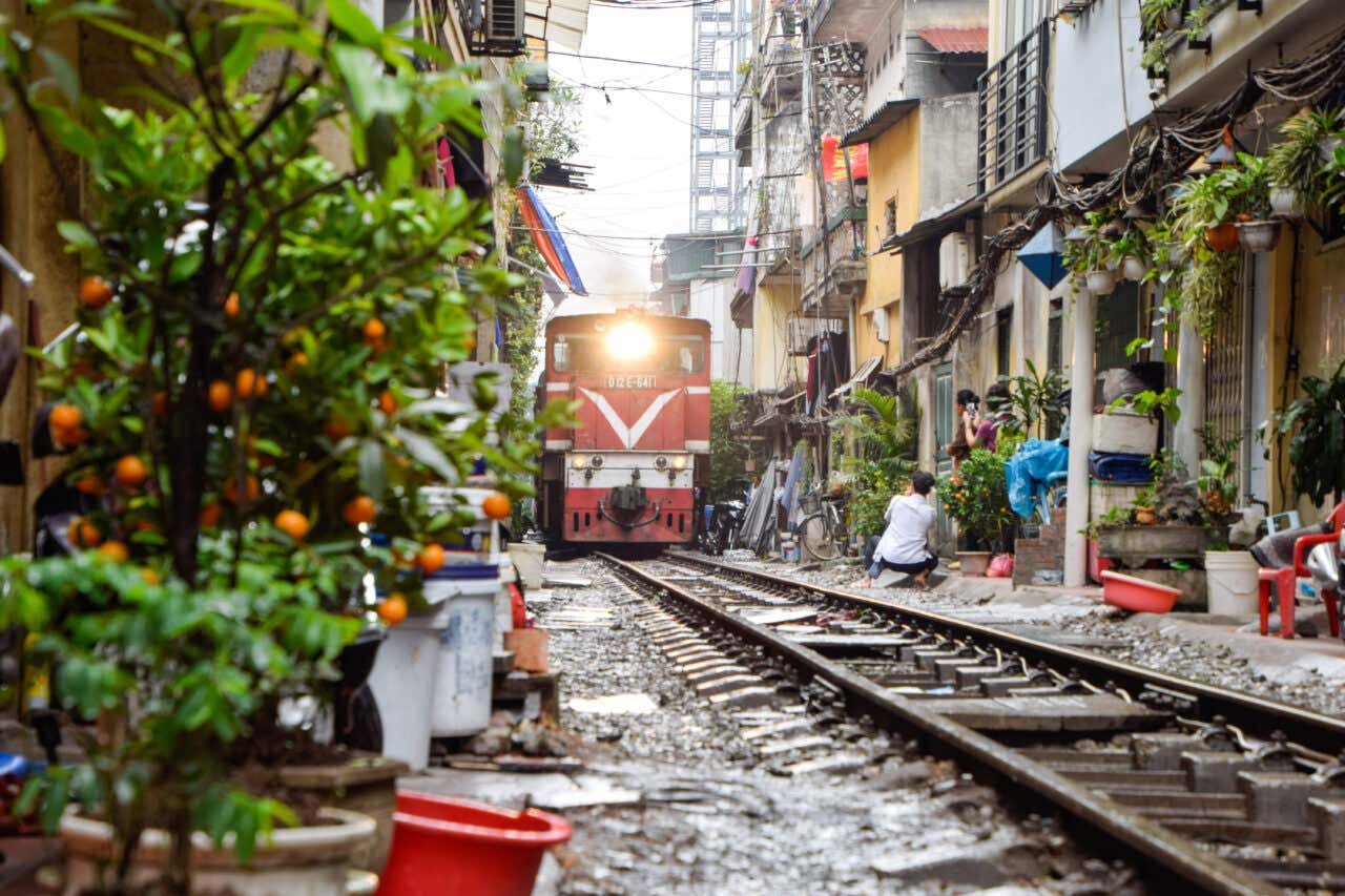 Treno che passa attraverso la stretta Train Street di Hanoi, con case e caffè a pochi centimetri dai binari, persone sedute lungo i lati e piante in vaso che decorano la via