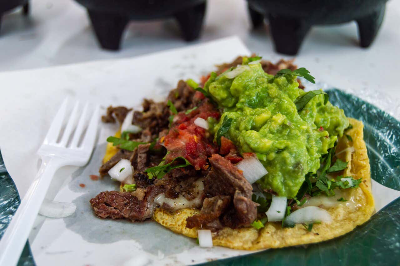 A vibrant, close-up food shot of a single, loaded taco or tostada on a plate, featuring grilled, chopped meat, diced onions, fresh cilantro, salsa, and a generous mound of bright green, chunky guacamole.