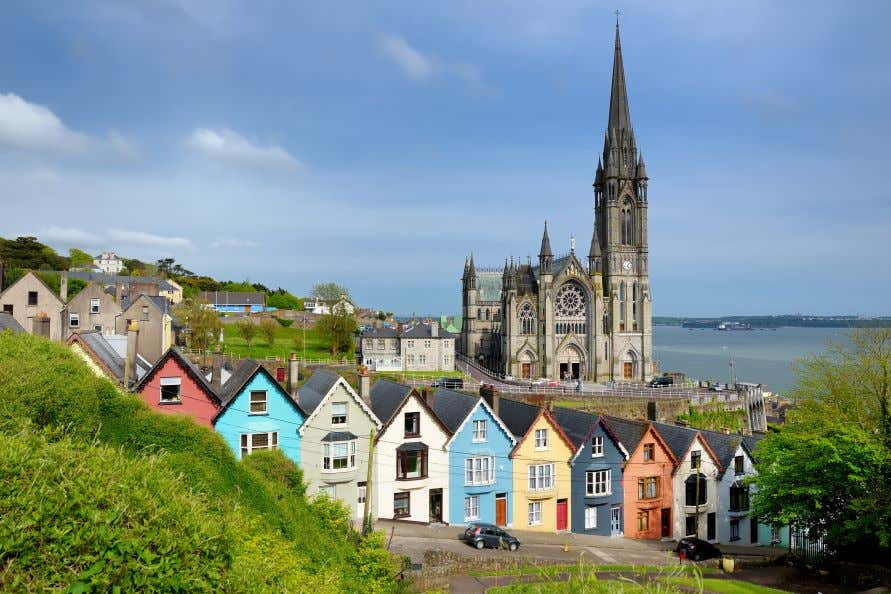 A view of St. Colman's Cathedral in Cobh towering over a seaside town with colorful houses on a hill.