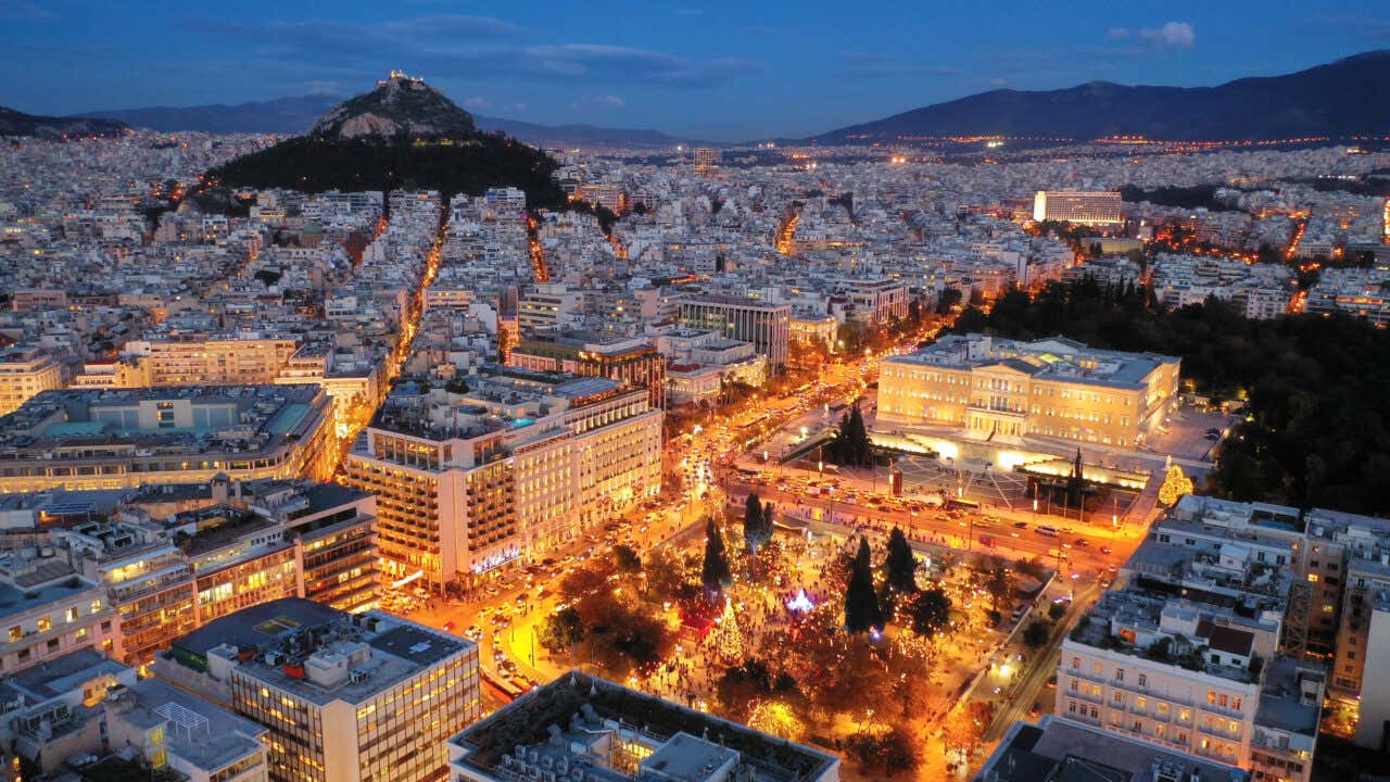 Atene vista dall'alto in una sera invernale, con una piazza al centro illuminata dalle luci di un mercatino di Natale e sullo sfondo la collina dell'Acropoli