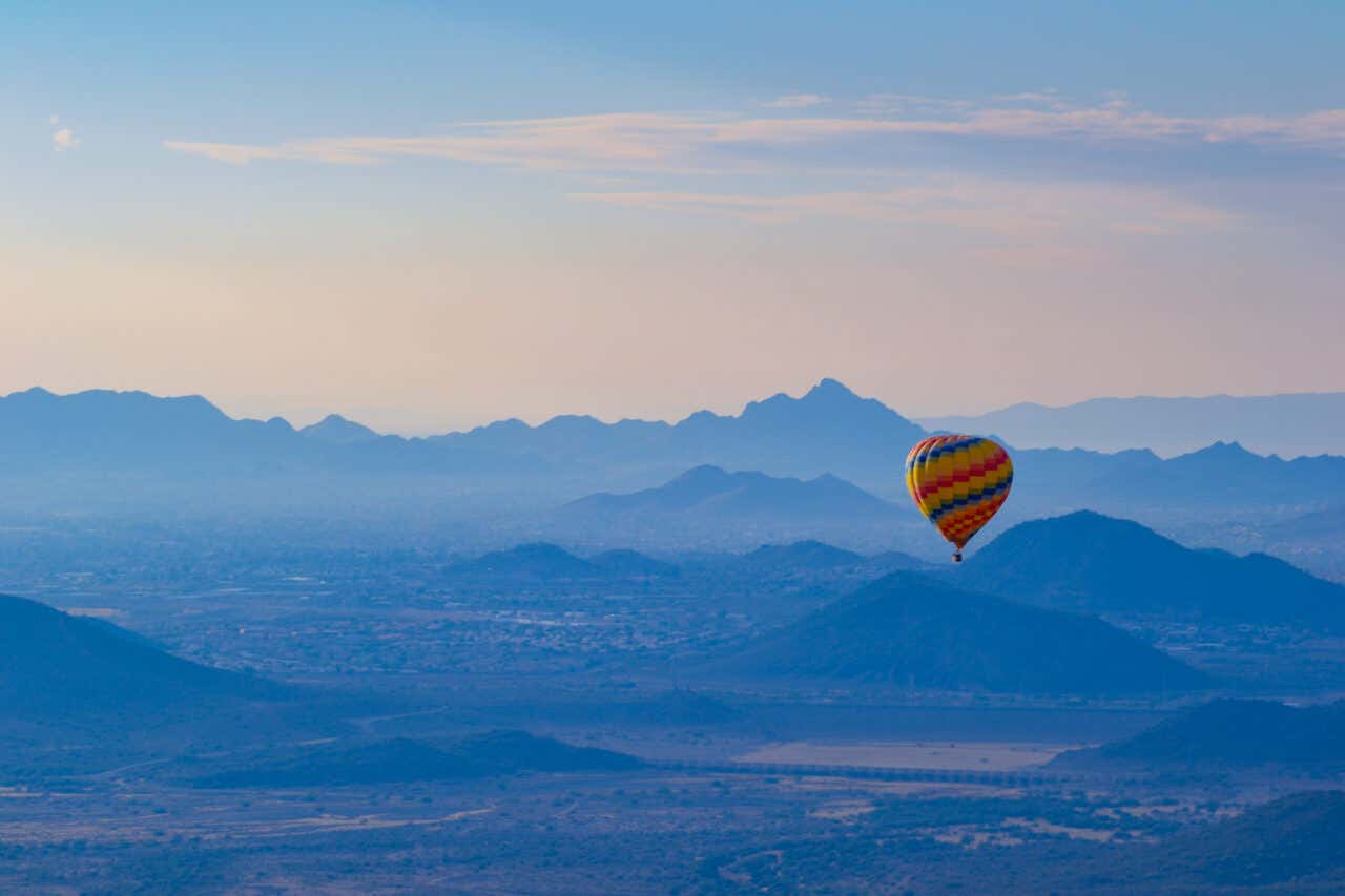 A colorful, striped hot air balloon floating high above a vast, hazy landscape of rolling, blue-tinted mountains and distant hills, set against a pale blue and white sky at sunrise.