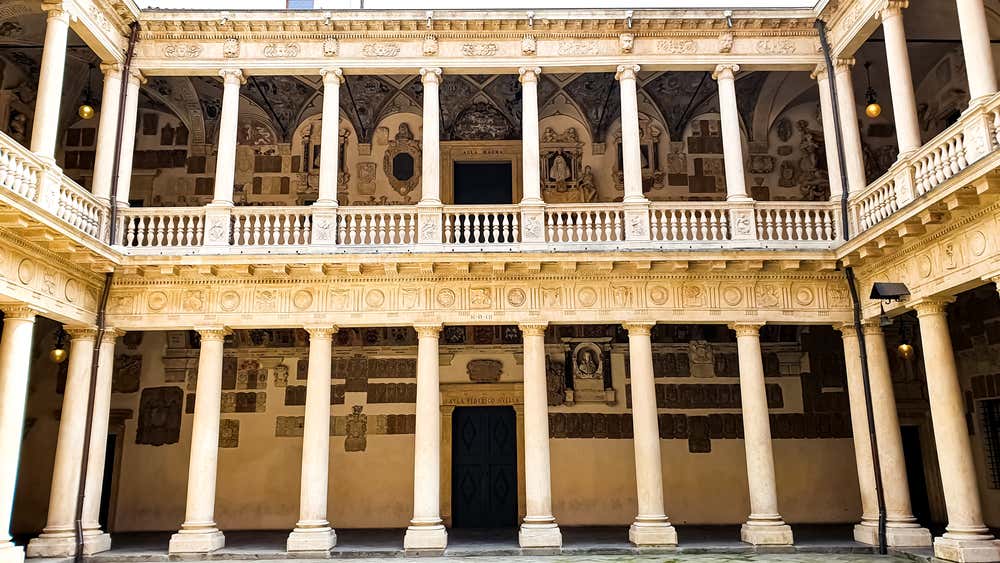 Vista su un lato del chiostro antico del Palazzo Bo: un cortile quadrangolare con doppio loggiato, colonne doriche al piano terra e colonne ioniche al piano superiore. Le volte del loggiato superiore sono decorate con stemmi araldici e dipinti.